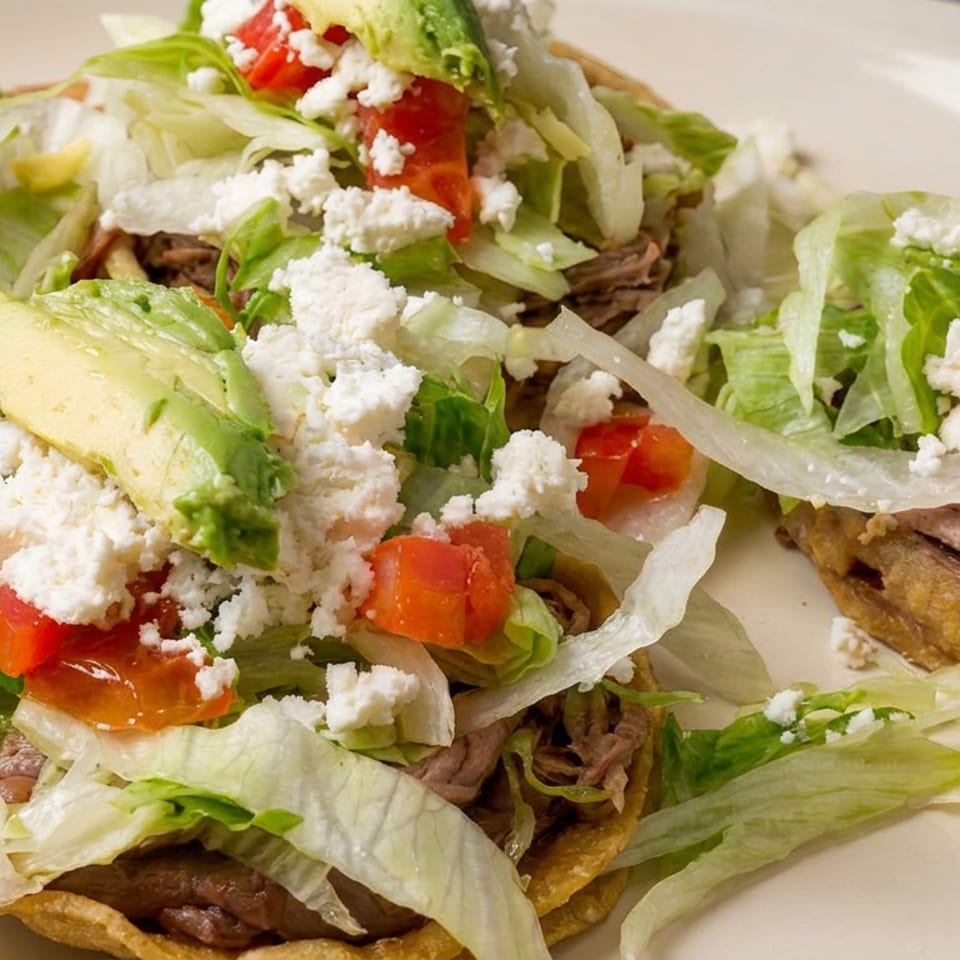 Tostadas topped with shredded beef, lettuce, tomatoes, onion, avocado, and crumbled white cheese on a plate.