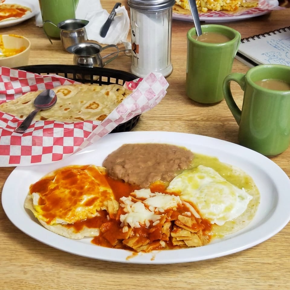 Mexican breakfast platter with eggs, beans, chilaquiles, tortillas, green mugs, and a diner sketching at the table.