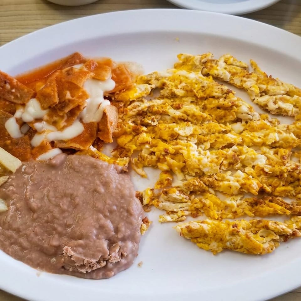 Plate of scrambled eggs, refried beans, fries, and sauced tortilla chips with cream, accompanied by tortillas and utensils.