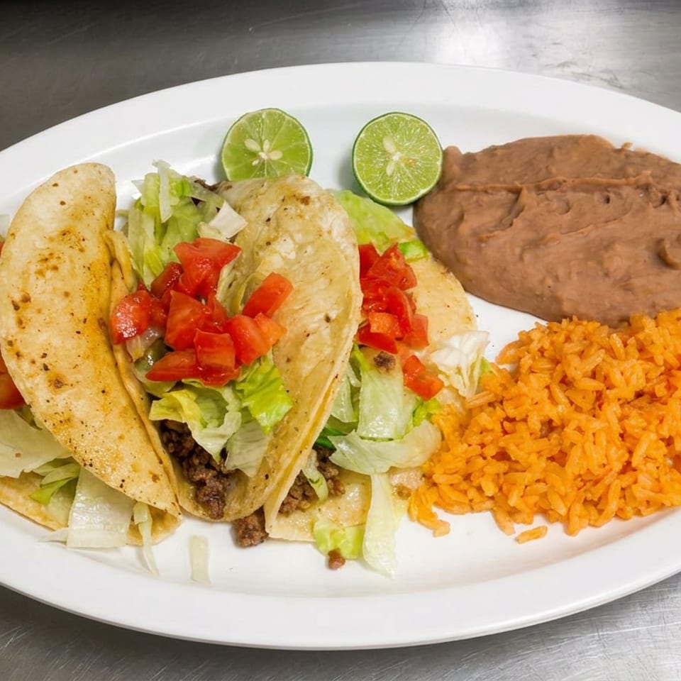 Plate of three tacos topped with lettuce and tomatoes, served with Mexican rice, refried beans, and lime wedges.